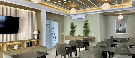 Una habitación de hotel moderna con televisor de pantalla plana, un sofá, una zona de comedor con mesas y sillas, y un techo con diseños geométricos.