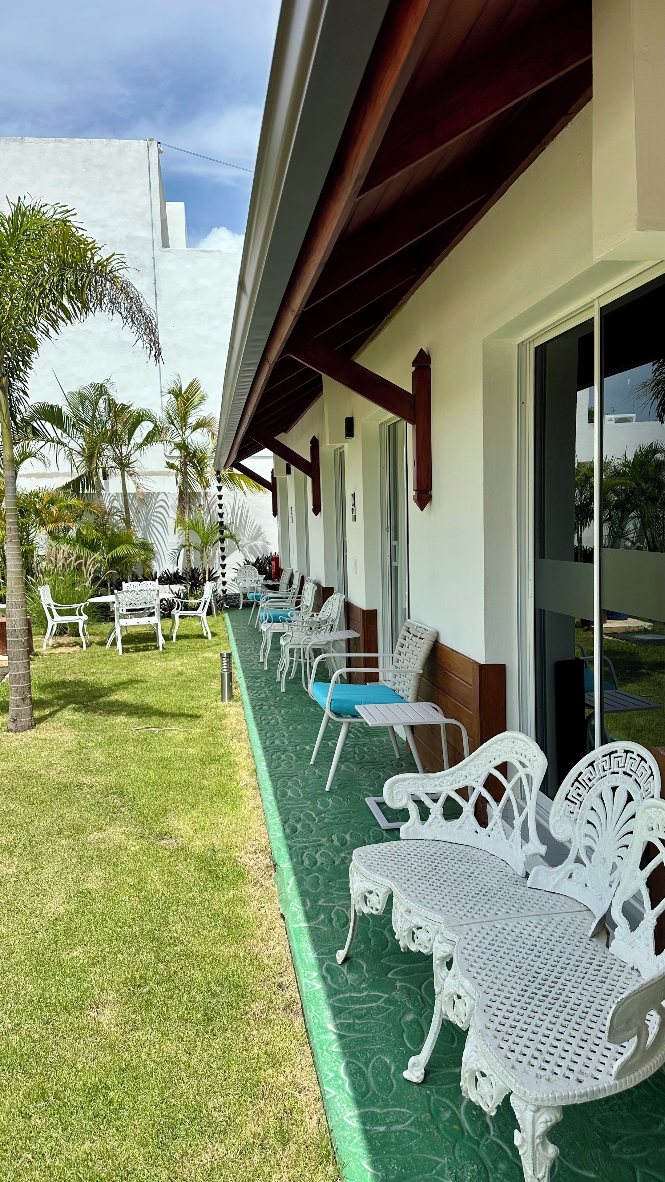 A covered outdoor seating area with white metal chairs and tables, a green mat, and palm trees.