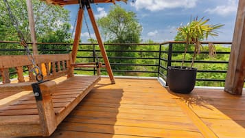 Una terraza de madera con un columpio, una planta en maceta y una barandilla con vistas a un paisaje verde exuberante.