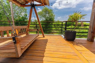 Une terrasse en bois avec une balançoire, un pot de fleurs et une rambarde donnant sur un paysage verdoyant et luxuriant.