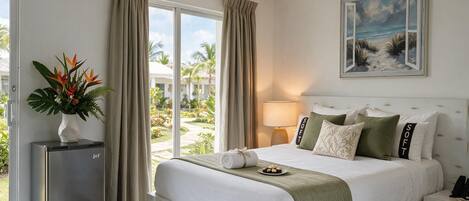 Una habitación de hotel con cama, mesita de noche, lámpara, planta y vistas al exterior.