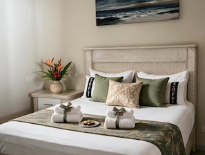Una cama bien hecha con una colcha de diseño verde y blanco, dos toallas dobladas cuidadosamente con un lazo verde, y un pequeño plato con aperitivos.