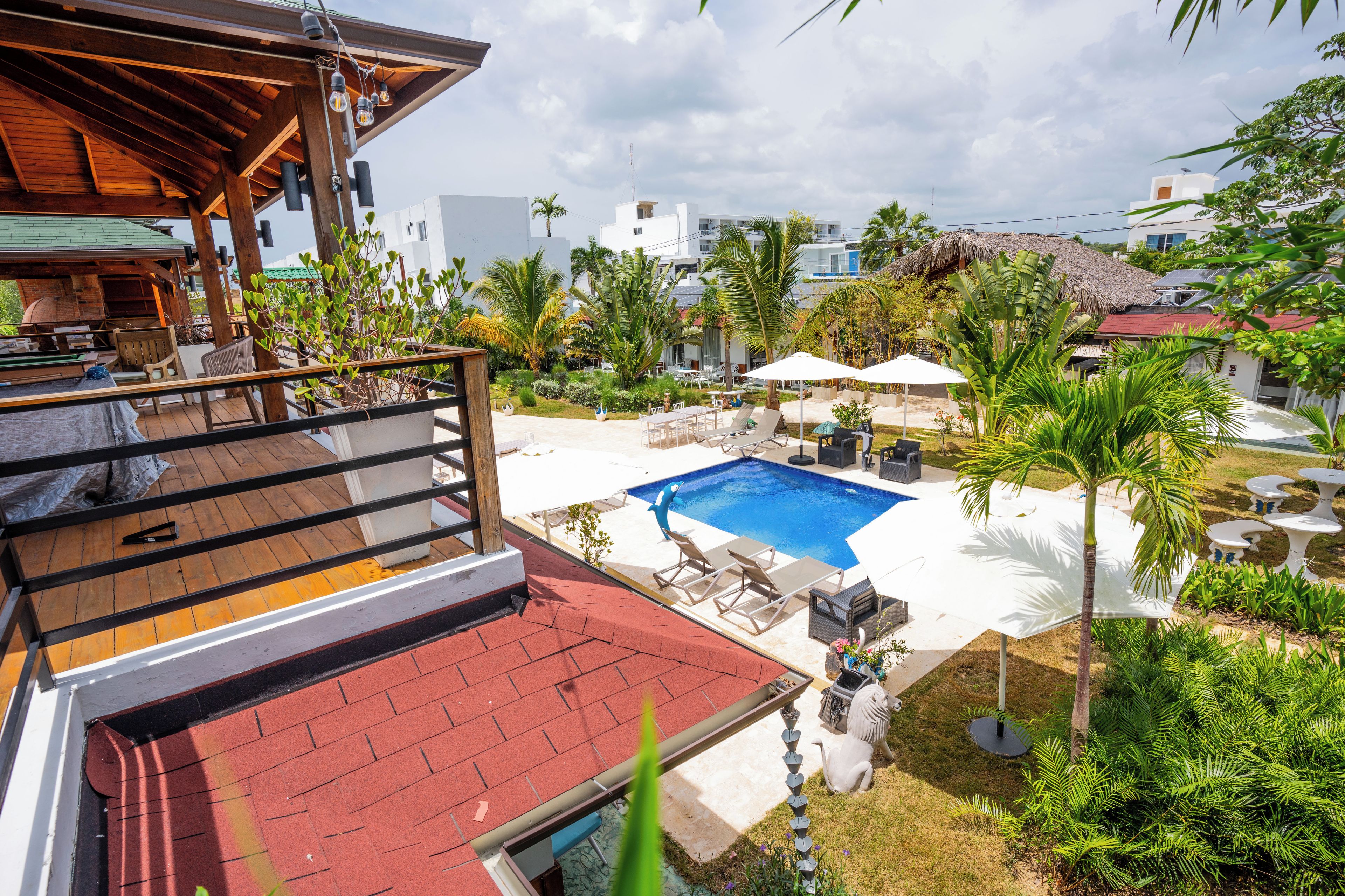 Área de piscina com espreguiçadeiras e guarda-sóis, cercada por vegetação e edifícios.
