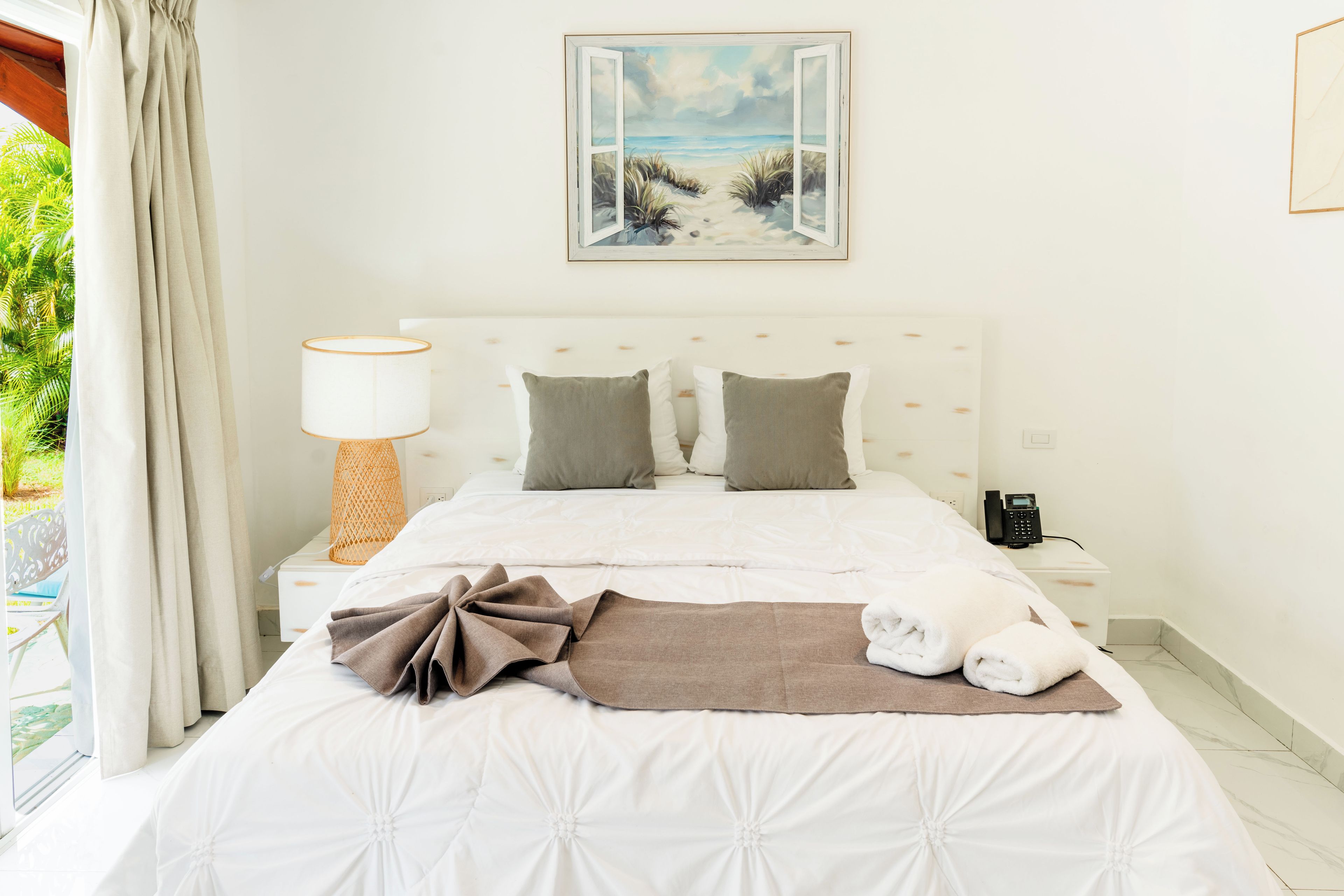 A hotel room with a bed, a bedside lamp, a painting, and a view of the outdoors.