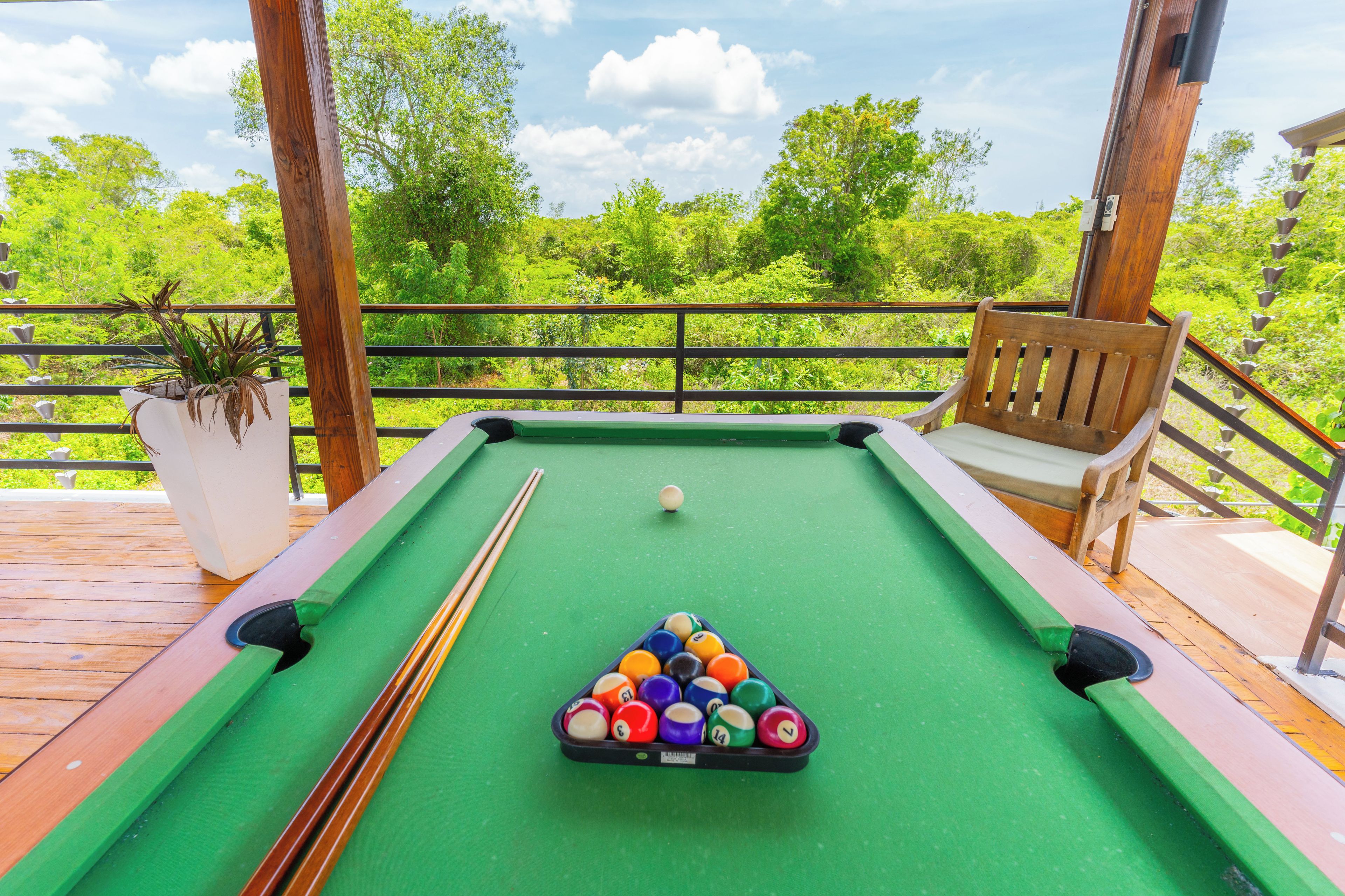 Mesa de bilhar com bolas e tacos em deck de madeira, com vista para vegetação.