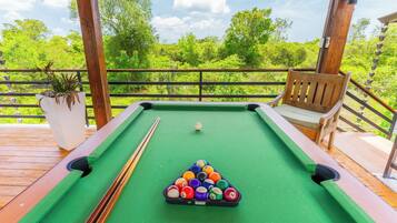 Mesa de bilhar com bolas e tacos em deck de madeira, com vista para vegetação.