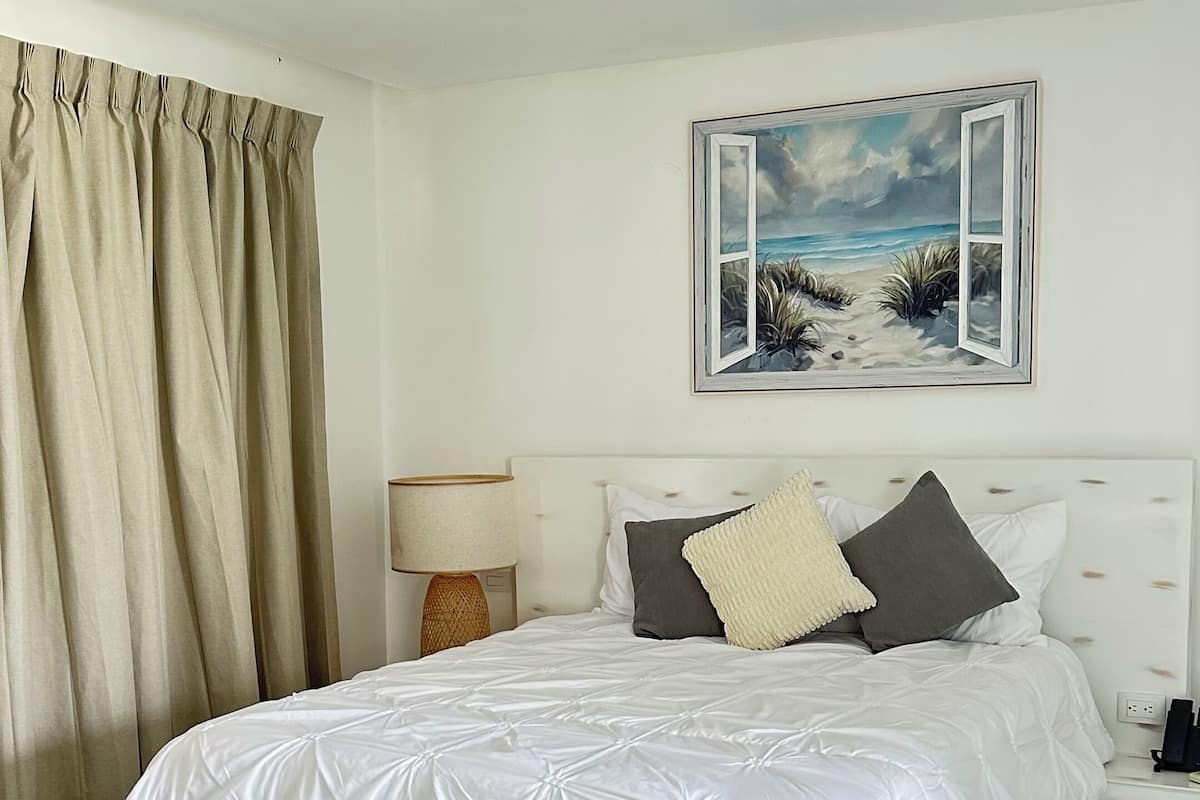 A bedroom with a bed, a nightstand, a lamp, a painting, and a window.