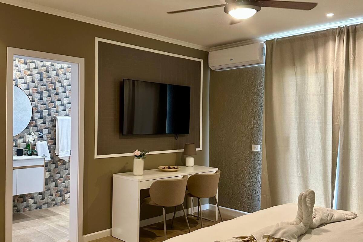 A modern hotel room with a bed, a desk with two chairs, a television, and an air conditioning unit.