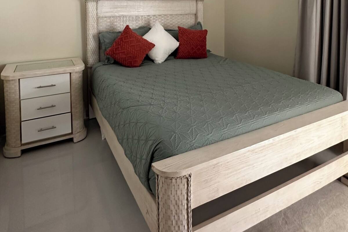 A bedroom with a bed, a nightstand, a drawer unit, a painting, and a curtain.