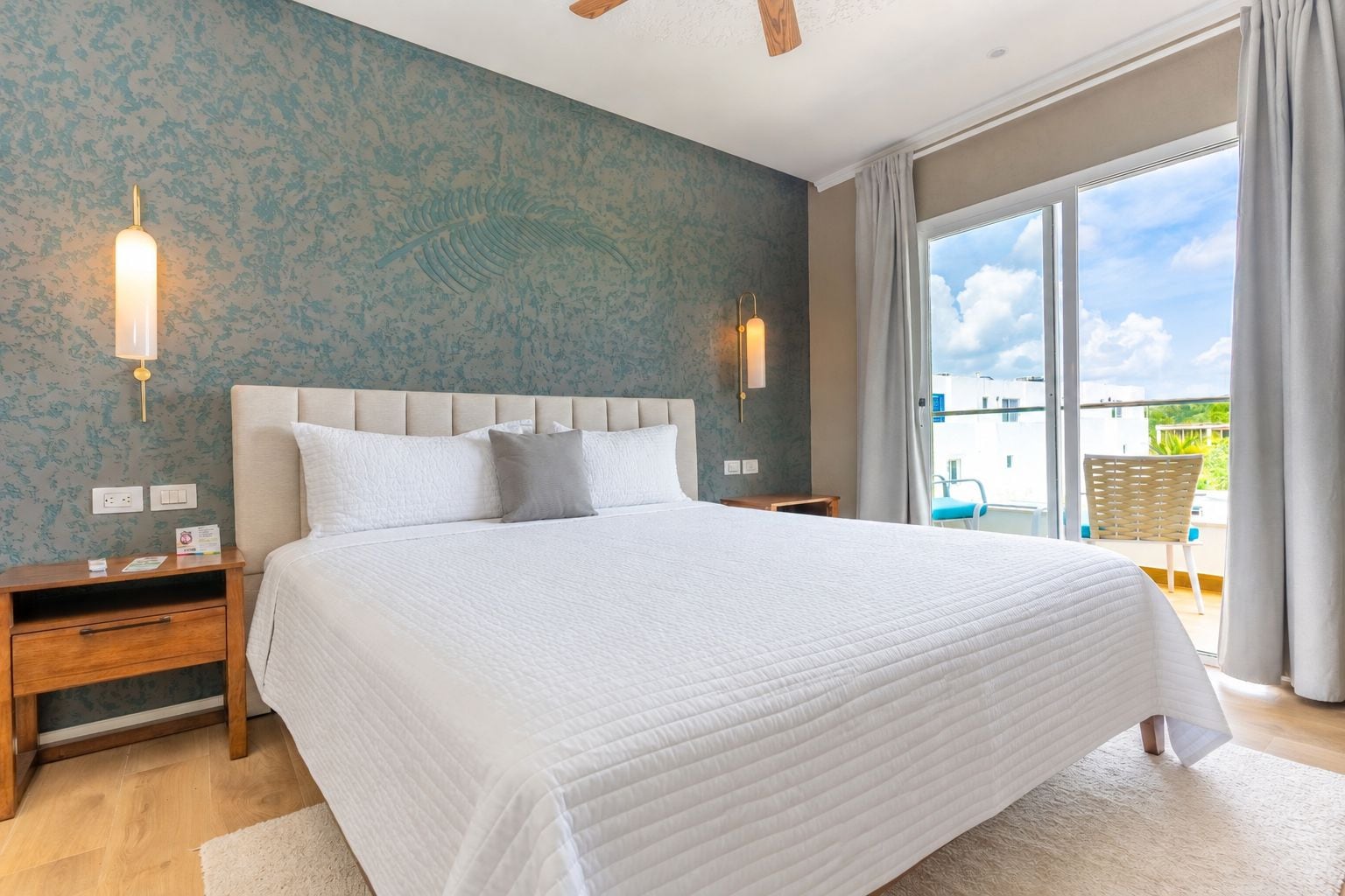 Quarto com cama grande, mesa de cabeceira, luminária fixada na parede e vista para o exterior por uma porta de correr de vidro.