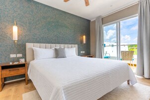 Quarto com cama grande, mesa de cabeceira, luminária fixada na parede e vista para o exterior por uma porta de correr de vidro.