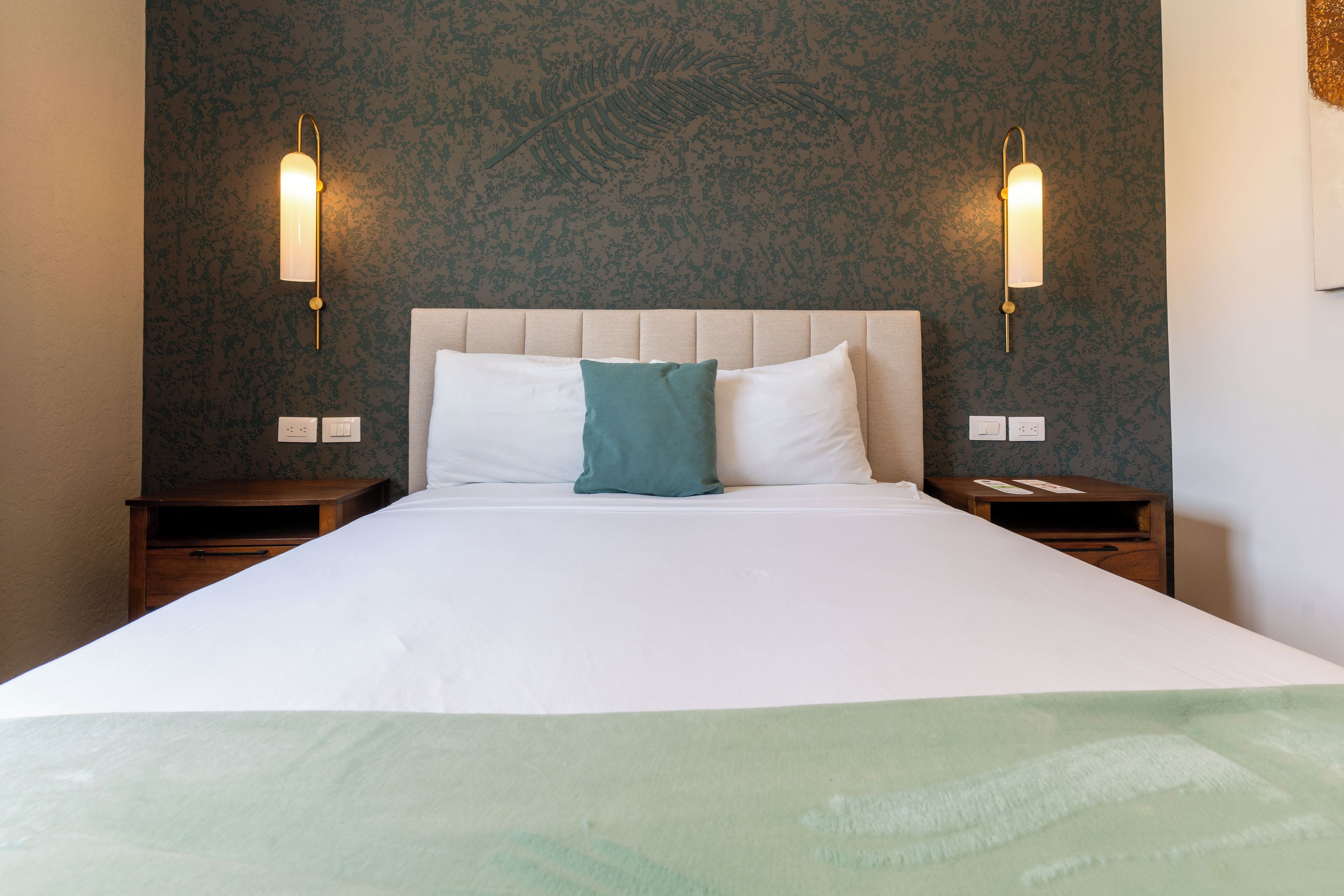 A neatly made bed with white linens and a teal pillow, flanked by two bedside tables with lamps, against a textured wall.