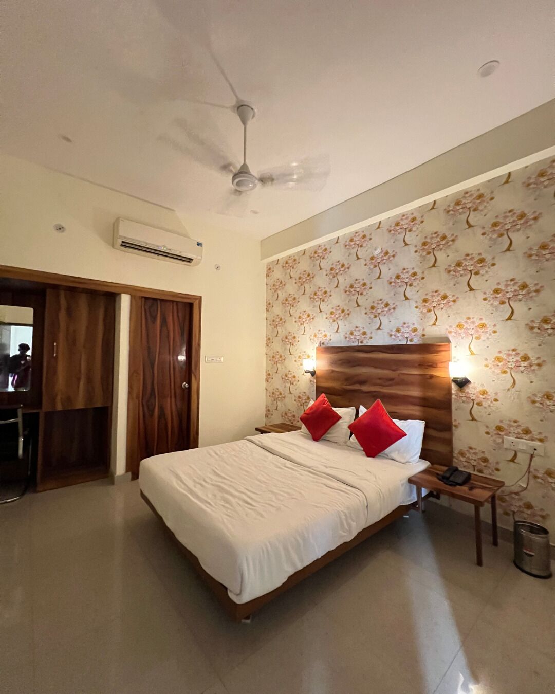 A hotel room with a bed, a wooden headboard, two bedside lamps, a nightstand, and an air conditioning unit.
