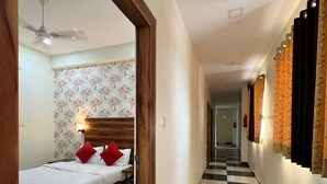 A hotel hallway with a checkered floor, a bed with red pillows, a floral wallpapered wall, and a wooden door frame with a number sign. - Hotel Shimla Grand (Lucknow)