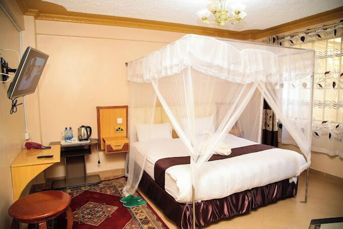 Habitación de hotel con cama bajo dosel, un escritorio con computadora, una silla y una pequeña mesa con hervidor.