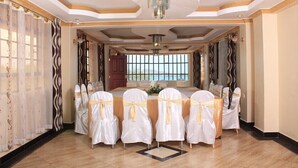 A banquet hall with a long table set for a formal event, featuring white chairs with gold sashes, a chandelier, and a view through the window. - Nuru Palace Hotel (Nakuru)