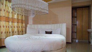 A round bed with a white headboard and multiple pillows, a lace chandelier, and a wooden wardrobe in the background. - Nuru Palace Hotel (Nakuru)