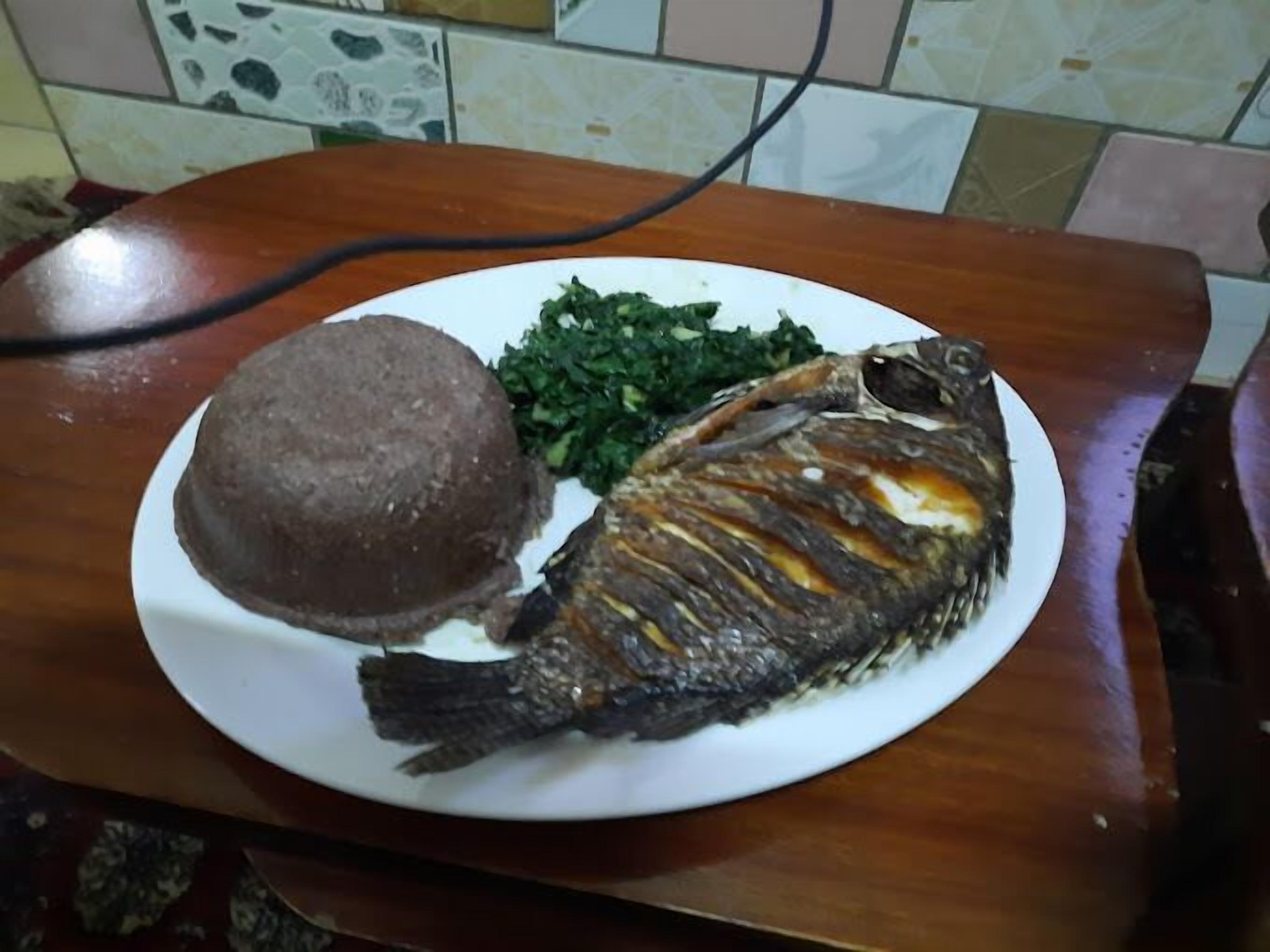 Un plato con un pescado frito entero, una porción de fufu y vegetales verdes.