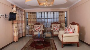 A living room with a sofa set, a coffee table, a TV mounted on the wall, and a large window with curtains. - Nuru Palace Hotel (Nakuru)