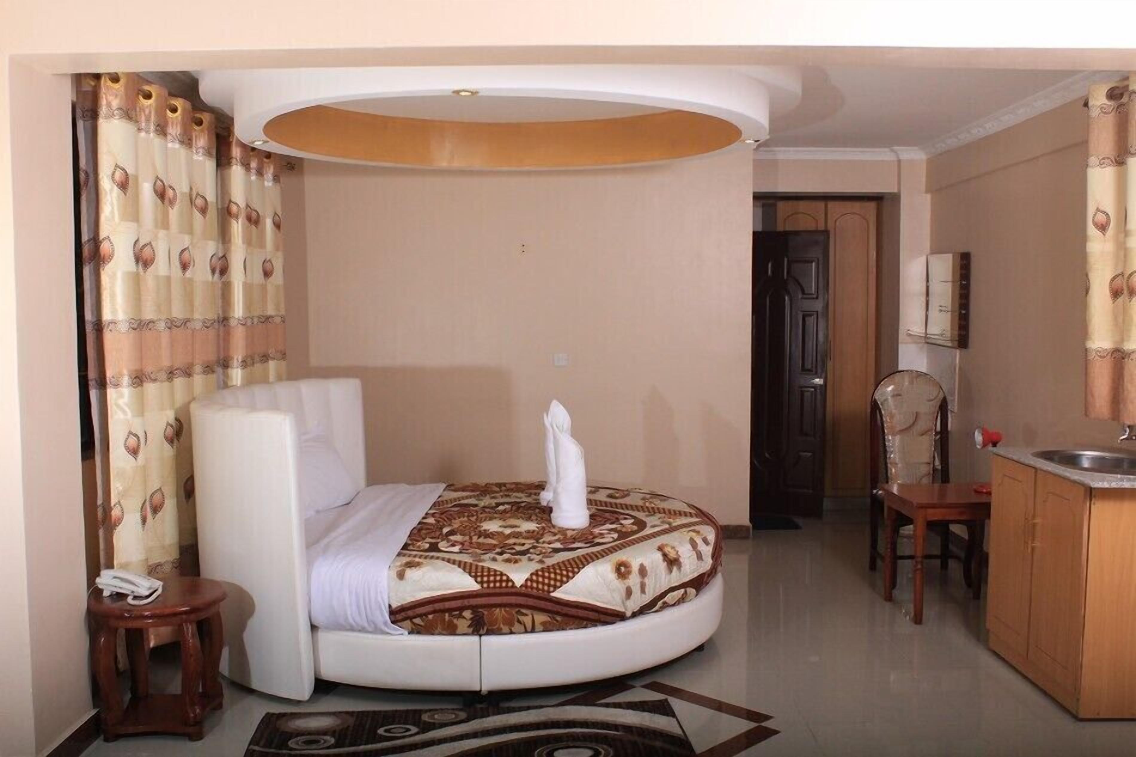 Una habitación de hotel compacta con cama, una mesita, una silla, kitchenette y puerta de baño.