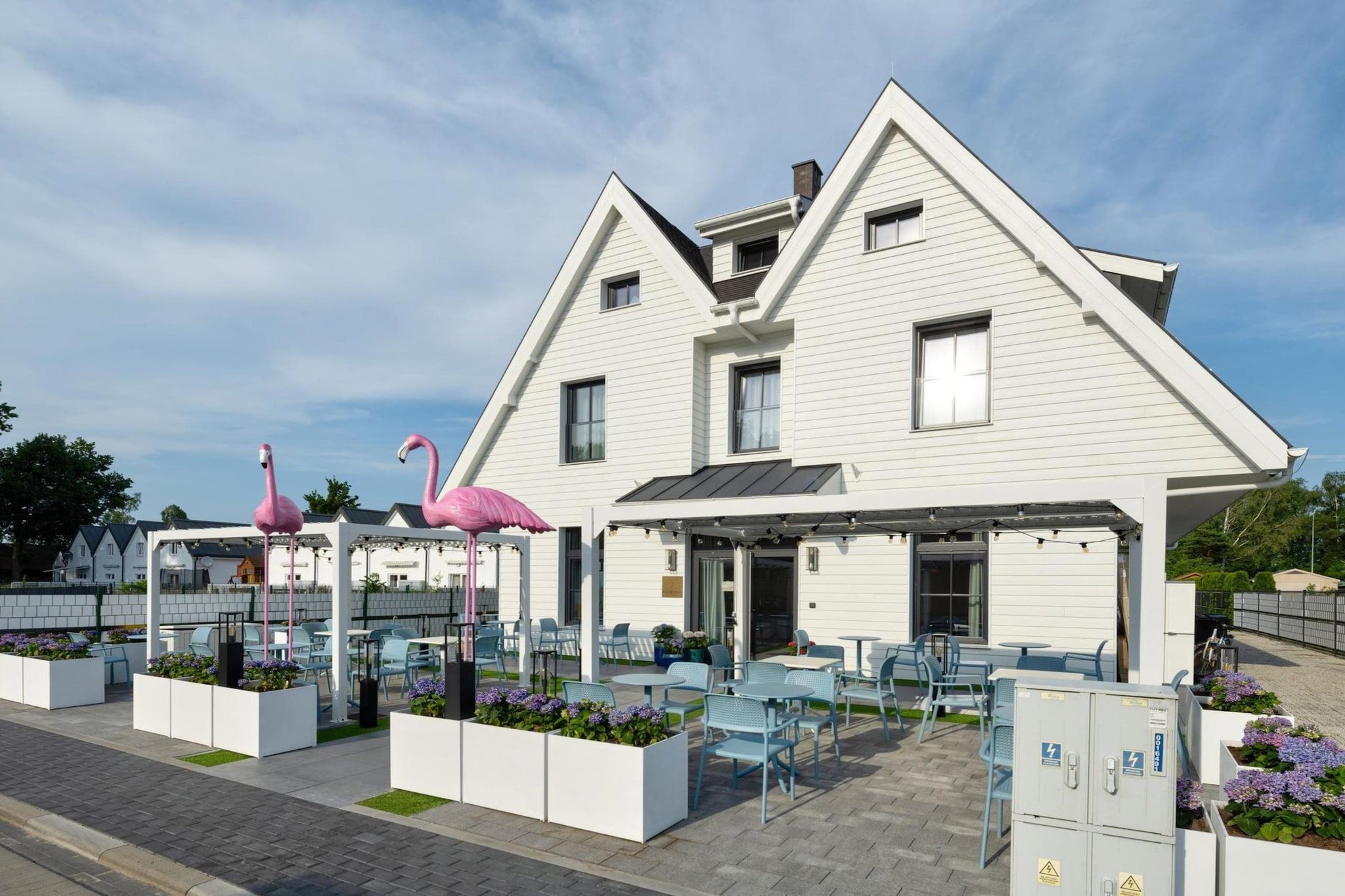 A white building with a sloped roof, outdoor seating area with tables and chairs, and pink flamingo statues.