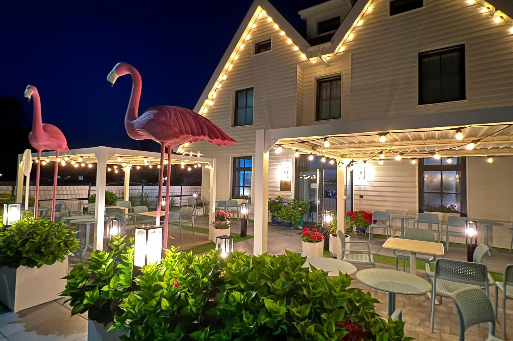 An outdoor patio area with tables and chairs, surrounded by potted plants and two large flamingo statues.