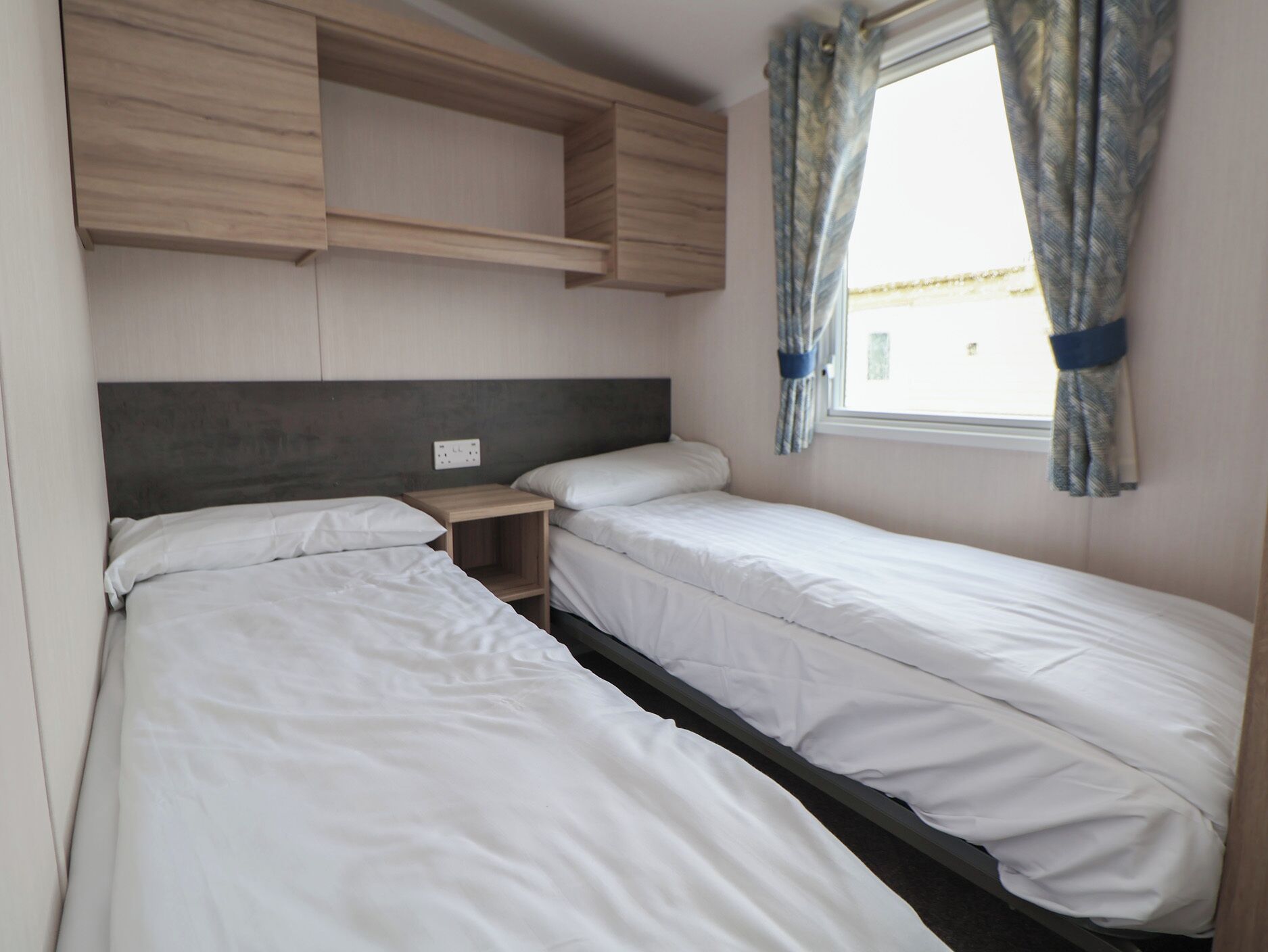 Une chambre à coucher de taille modeste, avec deux lits simples, une étagère en bois au-dessus et une fenêtre avec des rideaux.