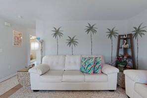 A living room with a white sofa, decorative pillows, a wall-mounted shelf with items, and a framed picture on the wall.