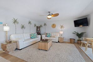 A modern living room with a white sofa, a coffee table, a ceiling fan, and a TV mounted on the wall.