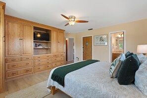 A bedroom with a large wooden wardrobe, a bed with multiple pillows, a ceiling fan, and a television on a shelf.