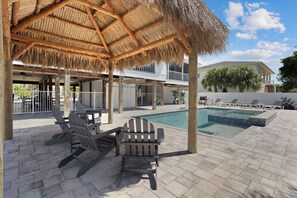 A pool area with a thatched roof, lounge chairs, and a view of a house.