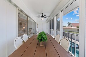 A covered outdoor area with a wooden table, chairs, a ceiling fan, and a view of a swimming pool.
