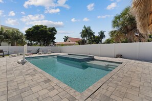Une piscine rectangulaire bordée de pierres, entourée d’une clôture blanche, et une terrasse blanche avec des chaises longues.