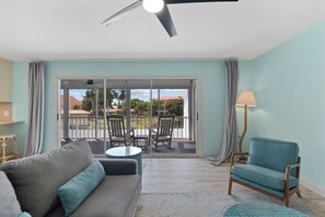 Un salon comprenant un canapé, un fauteuil, une table et une lampe. Une porte vitrée donne sur un espace extérieur avec une terrasse et des chaises.