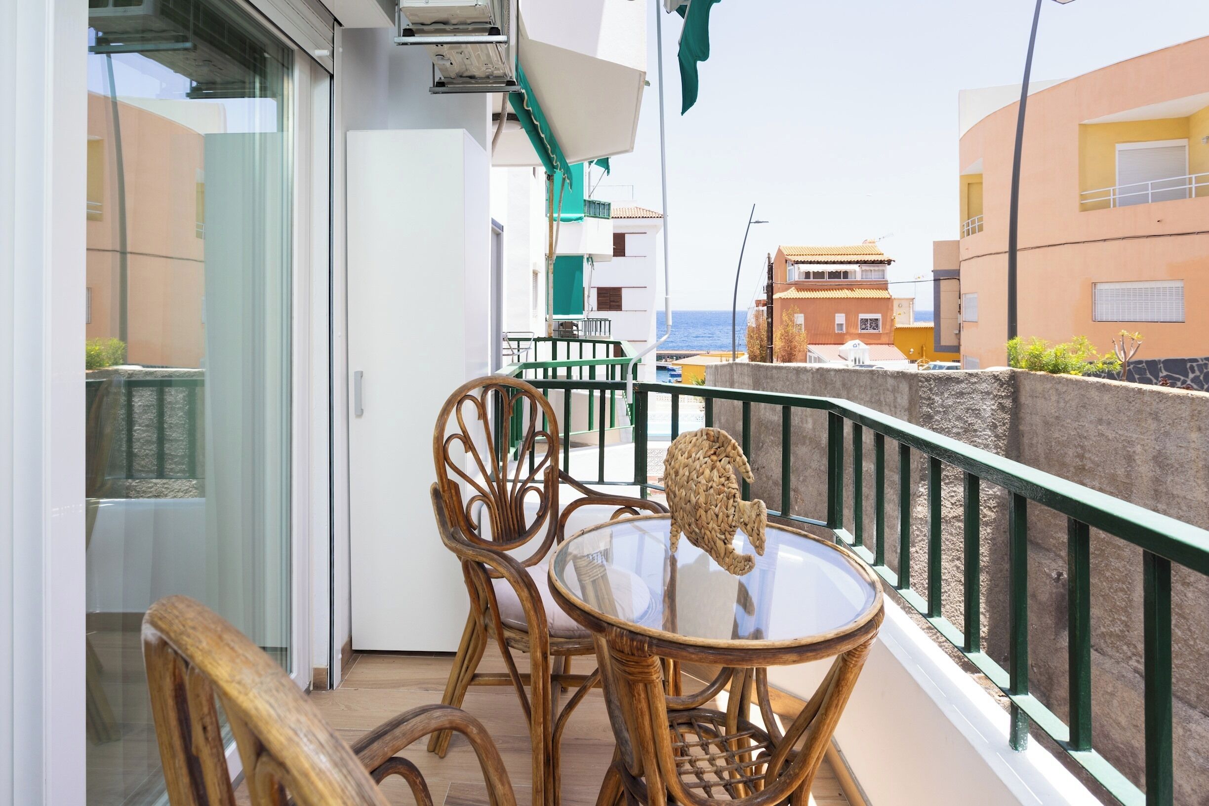 Ein Balkon mit Korbmöbeln und einem Glastisch, mit Blick auf eine Küstenlandschaft mit Gebäuden und dem Meer.