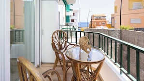 Un balcón con mobiliario de mimbre y una mesa de vidrio, con vistas a una zona costera con edificios y el mar.