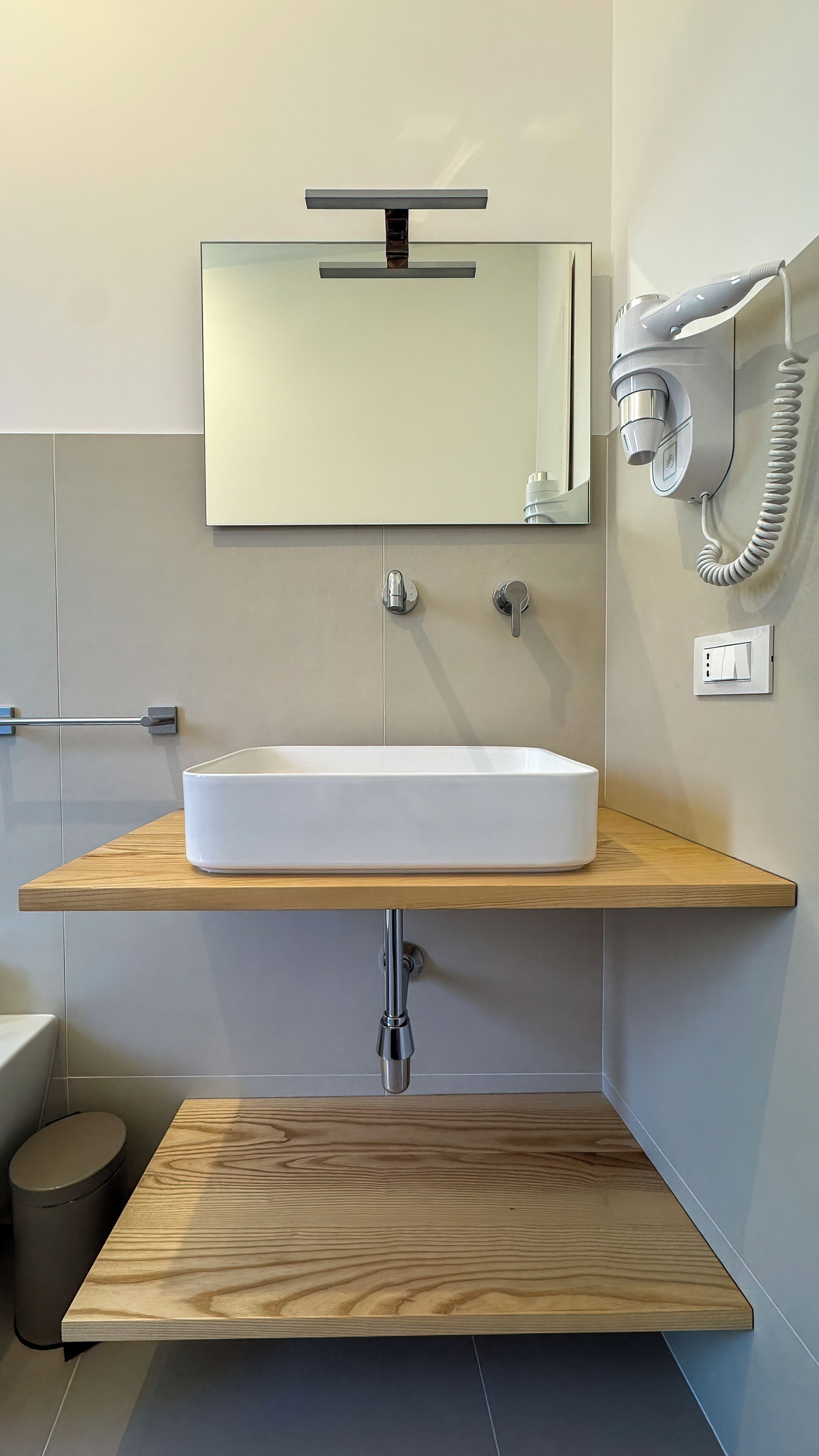 A modern bathroom with a rectangular sink, a mirror above it, a hairdryer mounted on the wall, and a toilet visible to the left.