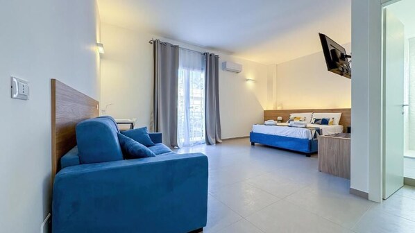 A modern hotel room with a blue sofa, a bed with a wooden headboard, a desk with a chair, and a TV mounted on the wall. - Tevergad Suite & Rooms (Terrasini)