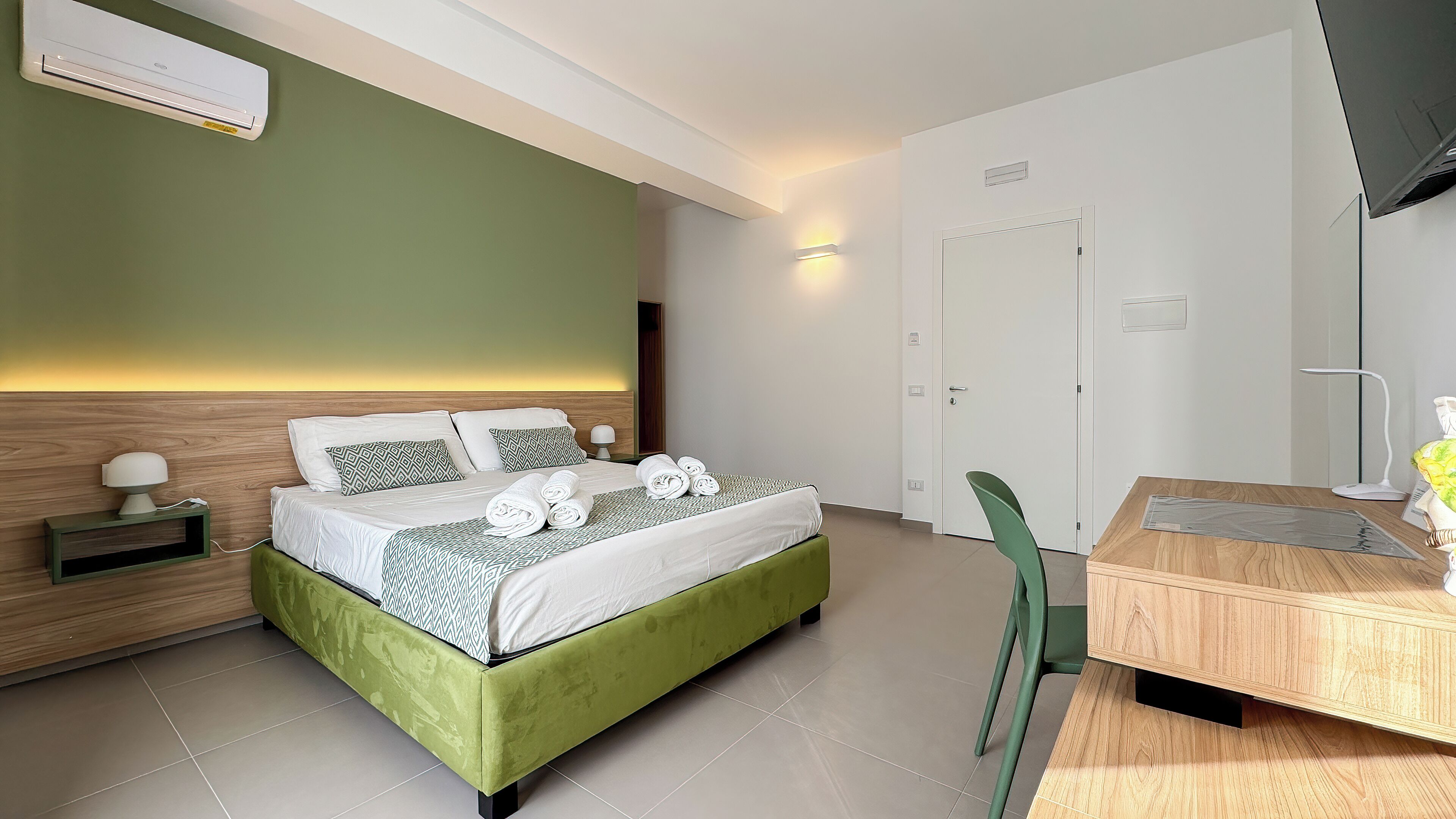 Habitación de hotel moderna con una cama tapizada de verde, una mesita de noche de madera, un escritorio con una silla y una puerta blanca.