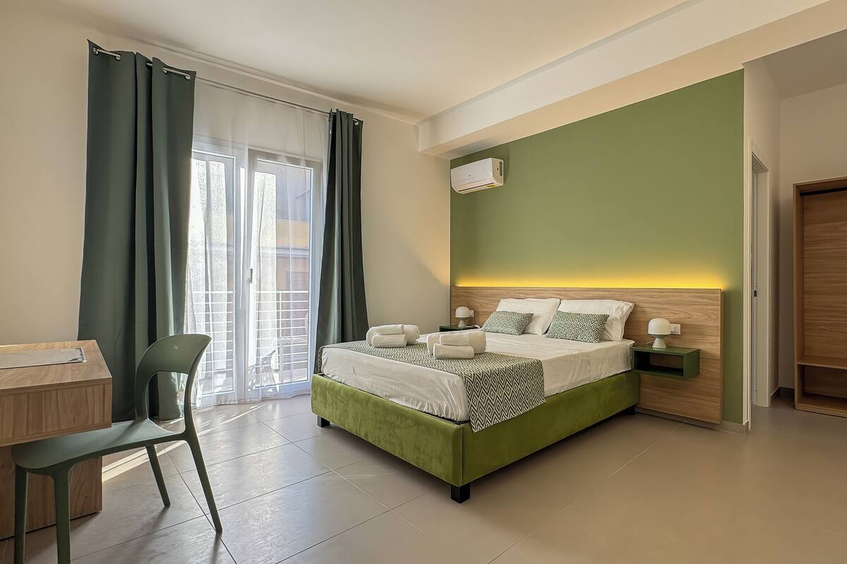 A modern hotel room with a green bed, a desk, a chair, and a sliding door to the balcony.