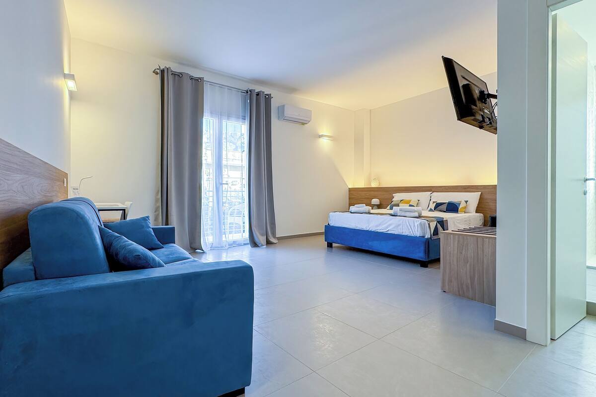A modern hotel room with a blue sofa, a bed with multiple pillows, a desk, and a TV mounted on the wall.