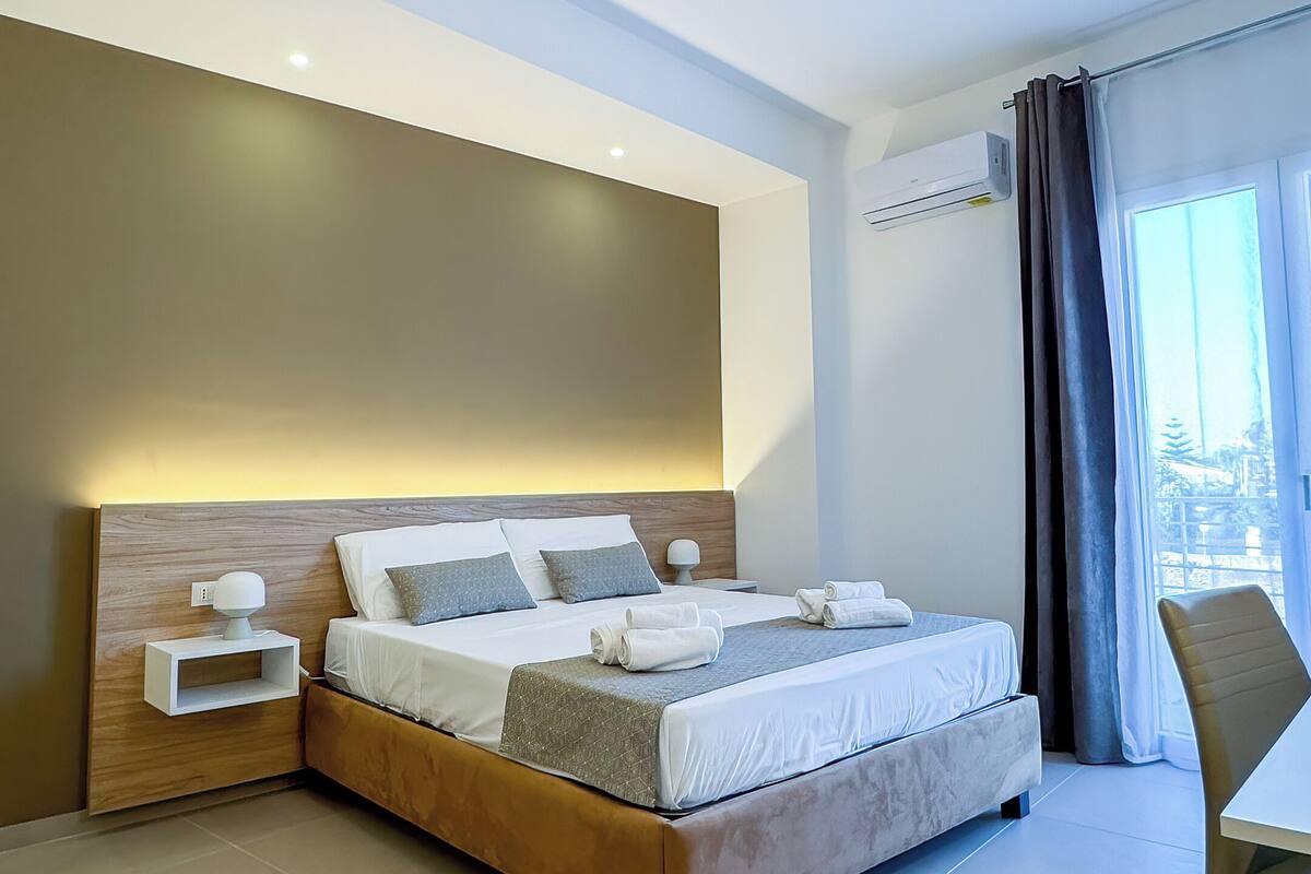 A modern hotel room with a large bed, bedside tables, a desk, and a view of the city through the window.