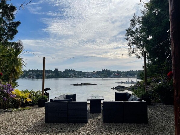 A patio with outdoor furniture overlooking a body of water with boats, surrounded by trees and a clear sky.
