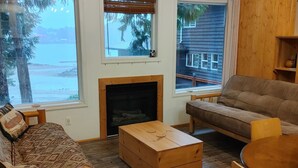 A cozy living room with a fireplace, a sofa, a coffee table, and a view of a wooden deck and house. - Ucluelet/Ocean/Treehouse Cabin - hot tub, kitchen, deck, Roku TV (Alberni Clayoquot Regional District)