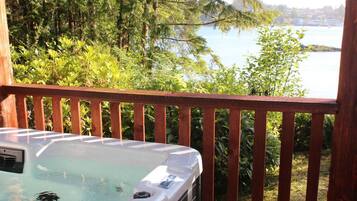 Relax in a hot tub on a deck overlooking a serene body of water surrounded by lush greenery.