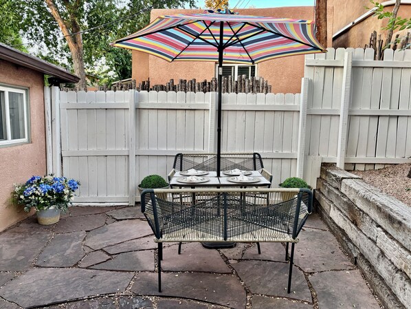 A patio with a table and chairs, a colorful umbrella, and a white picket fence. - La Casita Berger - Minutes away from Plaza and Canyon Rd (Santa Fe)