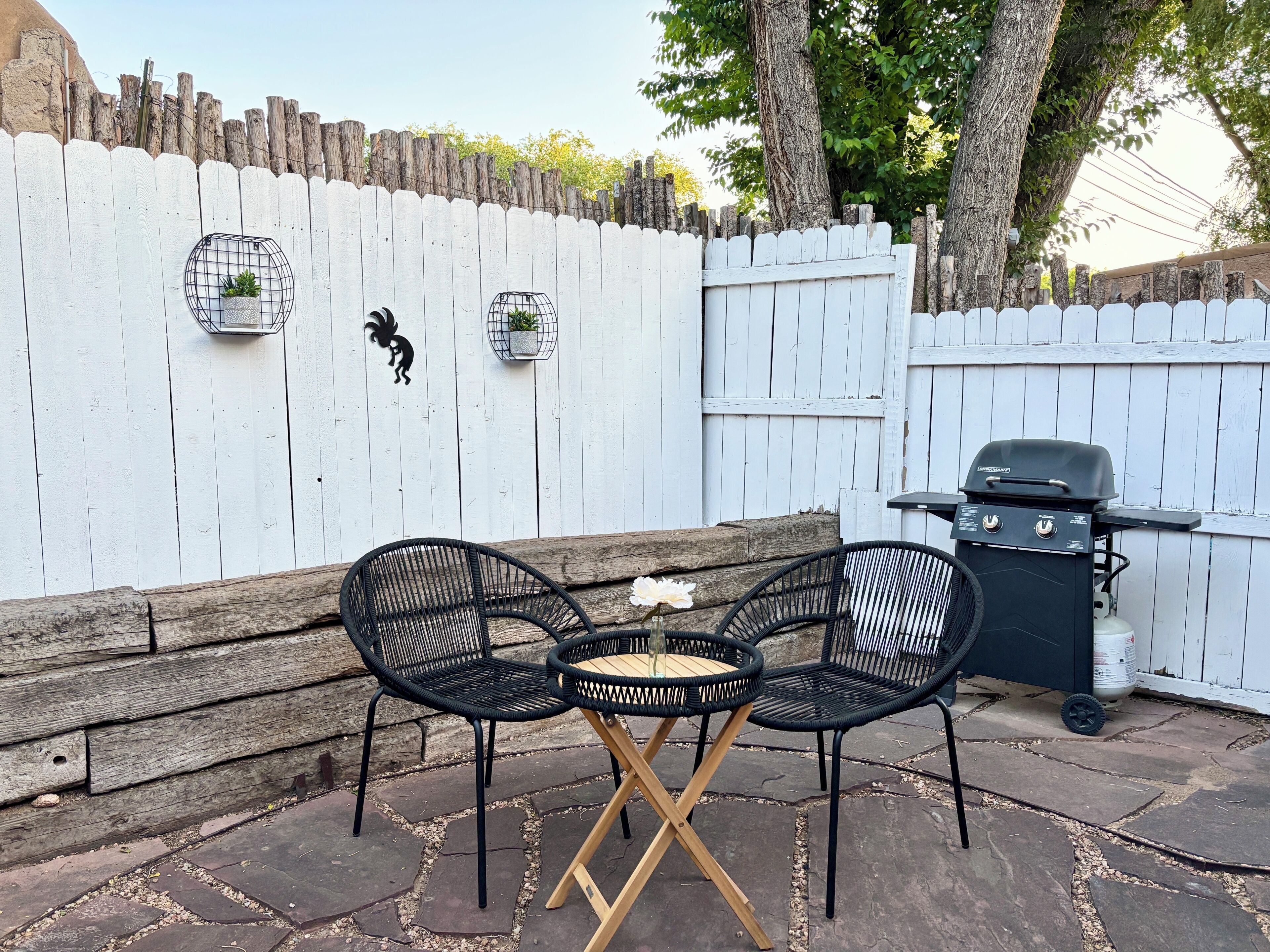 Een terras met twee zwarte wicker stoelen, een kleine ronde tafel, een barbecue en een witte picket hek.