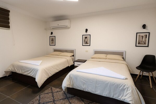 Habitación de hotel con dos camas individuales, una mesita de noche pequeña, una silla y un equipo de aire acondicionado en la pared.