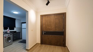 A hallway leading to a wooden door with a black handle, a washing machine, and a kitchen area with a microwave and a refrigerator.