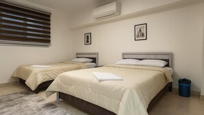 A hotel room with two beds, a window with blinds, a trash bin, and two framed pictures on the wall.